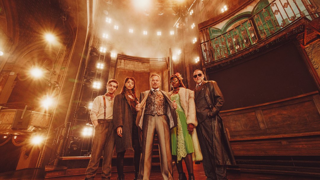 Elenco da montagem da Broadway de “Hadestown” no cenário do musical, com Perséfone de vestido verde e Hades de terno risca de giz